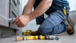 © Attasit - Close-up cropped view of plumber kneeling on bathroom floor, tightening pipe joint using professional tools, home repair setting.