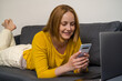 © Cavan - Cheerful woman laying down on sofa, holding a smartphone