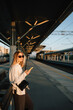 © Viktar Vysotski - Woman waiting for train at railway station during sunset, using her smartphone to check schedules and stay connected while enjoying golden hour light. Woman in casual style standing at railway station