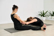 © TRAVELARIUM - Two young women practice partner yoga in studio. They sit on mats, stretching forward in synchronized pose. Light walls and plants create calming ambiance