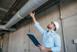 © Eliane - Confident construction engineer in hard hat inspecting industrial pipes indoors, holding a clipboard, demonstrating professional assessment, project management, and safety protocols in a building stru