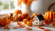 © Moonroad - Thanksgiving autumn decoration with small pumpkins and orange leaves surrounding miniature house on table near window creating warm and cozy seasonal atmosphere