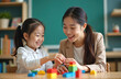 © Maryna - Smiling Asian teacher interacts with young girl with Down syndrome playing with colorful building blocks. Classroom setting with books, chalkboard creates atmosphere of learning, development. Focus