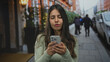 © Krakenimages.com - Young hispanic woman using smartphone outdoors on a busy street, focused on her phone amidst pedestrians and urban decor, capturing a modern city lifestyle scene.