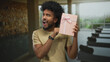 © Krakenimages.com - Man happily holding a pink gift box indoors at a resort pool area, expressing joy and surprise with lush greenery visible through large windows.