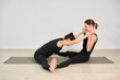 © TRAVELARIUM - Two adult women perform partner yoga pose on mat in minimalist studio. One is leaning forward, and other supports her, creating sense of connection and balance
