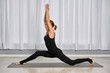 © TRAVELARIUM - Adult woman with fair skin performs lunge yoga pose on mat indoors. Side view highlights her concentration and flexibility against serene curtain backdrop