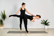 © TRAVELARIUM - Female instructor helps woman in black perform yoga pose on mat in serene studio. Natural light, light walls, and plants create calm ambiance
