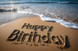 © Monique - Happy Birthday carved in smooth sand on an empty beach and waves gently lapping at its edges.