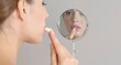 © Richafuji - A profile view of a woman applying lip balm in front of a mirror on a gray background. The woman has fair skin and is focused on the reflection in the mirror.