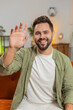 © Andrii Iemelianenko - Happy young Caucasian man smiling friendly at camera and waving hand gesturing hello, hi, greeting talking while sitting on sofa in living room at home. Happy bearded guy recording vlog at home.