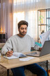 © Andrii Iemelianenko - Young Indian man freelancer sitting at table reviewing financial reports on laptop, holding documents and receipts, taking notes in notebook. Hispanic guy sits nearby at home, focused on his own task.