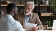 © Johannes - Serious focused mature businessman employer hr or client holding cv or contract listening manager, applicant during job interview, two diverse ethnic partners having negotiations at office meeting.,