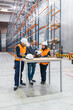 © Koldo_Studio - Engineers and manager wearing hardhats discussing logistics over blueprints in warehouse with tall shelves