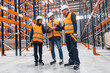© Koldo_Studio - Warehouse workers and manager wearing safety vests and hardhats discussing logistics using digital tablet and barcode scanner in large distribution warehouse