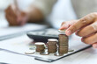 © Wanan - Concept of Finance. Budgeting. Taxes. Accounting, Investment. Businessman stacking coins and doing financial calculations on a desk with documents and a computer.