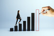 © Who is Danny - Businesswoman climbing bar graph steps as hand draws final growth column on blue background representing success and corporate progress.
