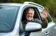 © Viktor - Senior man in car smiles, waves hand in friendly greeting. Happy driver at window, smiling face, looks at you. Elderly man enjoying drive. Active retired person. Travel concept.