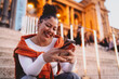 © BullRun - young woman enjoys online shopping while relaxing on historic city steps. Female holds credit card and smartphone, bright smile reflecting satisfaction with digital convenience and financial solutions