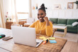 © BullRun - Smiling young Black woman working remotely with laptop and earphones, enjoying positive video call in cozy home interior with sticky notes and stationery on wooden desk