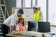 © AU USAnakul - Group of engineers wearing safety helmets and vests analyzing building plans on computer in modern office, showing teamwork and precision in construction.
