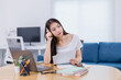 © Tj - Young Asian businesswoman working from home, sitting at a desk with a laptop and documents, holding a pen, gazing thoughtfully into the distance while contemplating her next move