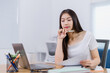 © Tj - Young Asian businesswoman holding a pen and reading documents while working on a laptop, concentrating on project strategy and analyzing reports at an office desk