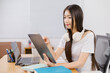 © Tj - Young Asian businesswoman using digital tablet and working at home office, headphones around her neck, laptop and notebooks on the desk, working or studying online concept