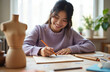 © Vadym - Asian woman artist draws with crayon on paper at home table. Young girl smiles creating artwork with wax crayons. Focused female artist pursues creative hobby indoors, enjoying leisure time.