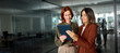 © insta_photos - Two happy busy professional business women, diverse female employees workers talking using digital tablet device working together in partnership while standing at work in office. Candid photo.