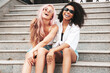© halayalex - Two young beautiful smiling hipster female in trendy summer clothes.Sexy carefree multiracial women posing on the street background.Positive models having fun in sunglasses. Sitting at the stairs