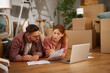 © Stockphotodirectors - A couple is lying on the floor in a welcoming room, discussing moving details while reviewing paperwork on a laptop. Cardboard boxes are scattered around, indicating the transition to their new home.