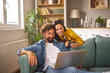 © Stockphotodirectors - A man and woman are sitting together on a green couch, with the man focused on a laptop while the woman smiles and holds a cup. Sunlight streams into the cozy living room.