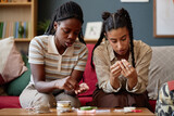 Black young adult woman and her girlfriend sitting together making handmade jewelry using beads and string, both focused on crafting with hands close to materials