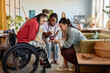© Seventyfour - Three young adult women, including Black and Caucasian, gathering in art studio, smiling and interacting with smartphone, one woman in wheelchair with disability participating in conversation