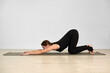 © TRAVELARIUM - Young woman with light skin practices extended puppy pose on gray yoga mat. Minimalist studio and soft lighting create serene ambiance, emphasizing relaxation and focus