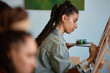 © Seventyfour - Young adult multicultural woman painting on canvas in art studio, holding palette and brush, focusing on creative process, long braided hair visible, other people blurred in foreground