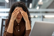 © Liubomir - A woman stressed out at her desk, touching her head, looking down, with a laptop in front of her in an office setting.