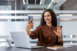 © Liubomir - A young woman in a modern office is smiling while holding her phone, possibly video conferencing. She's waving at the camera.