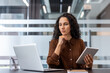 © Liubomir - A woman in a modern office contemplates while holding a tablet and using a laptop, surrounded by glass walls.