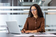 © Liubomir - A woman with curly hair and glasses works on a laptop in a modern office setting, focused and professional.