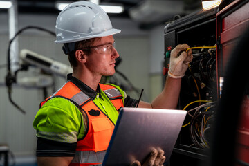 A man in a green and orange vest is working on a laptop. Electrical engineer or technician using a laptop for diagnostics and maintenance of a machine control panel in a factory.