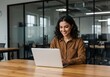 © shahida  - Smiling young woman with curly dark hair working on a laptop at a wooden desk in a modern office