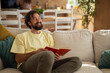 © Dexon Dee - Smiling man sitting on a sofa and reading a book at home, enjoying a peaceful and relaxing moment in the living room.