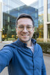 © Liubomir - A man wearing glasses takes a selfie in front of a modern glass building, smiling towards the camera.