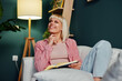 © djile - Smiling woman relaxing on sofa reading book and daydreaming in living room