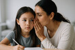 © Sandor - Mother whispering secret to curious daughter as she writes, capturing intimate family bond and trust in a cozy home setting
