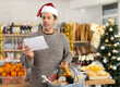 © JackF - Focused man in festive hat and casualwear looking at shopping list while moving along aisle between displays and holding cart with