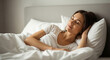 © yollieee - Young woman peacefully waking up in bed, smiling with eyes closed, wrapped in white sheets. Morning light and relaxed atmosphere.