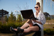 © keleny - Woman working on a laptop outdoors in a stylish outfit on a sunny day in a park setting